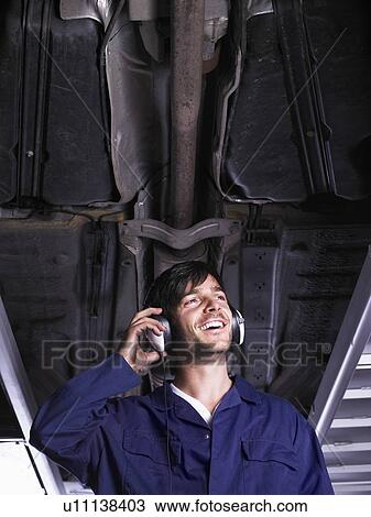Stock Image - Mechanic standing under raised car listening to music. Fotosearch