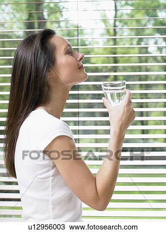 Woman with eyes closed standing by a window holding a glass of water. View Large Photo Image Stock Image - Woman with eyes closed standing by a window holding a glass of water.. Fotosearch