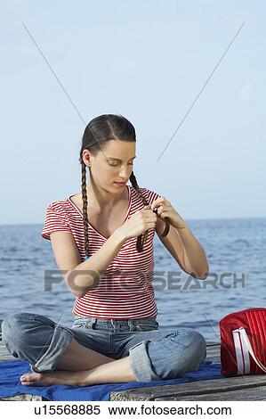 Stock Photography - Young girl sitting on jetty plaiting herself pigtails.. Fotosearch