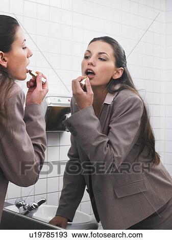 Stock Image - Woman reflected in office washroom mirror applying make-up. Fotosearch