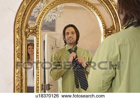 Stock Photograph - young man adjusting tie in mirror watched by woman. Fotosearch