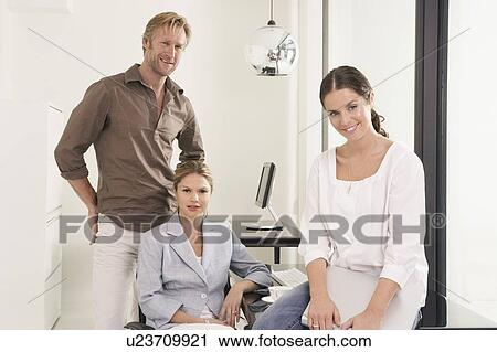 Stock Image - Man and two women in office woman seated on desk.. Fotosearch