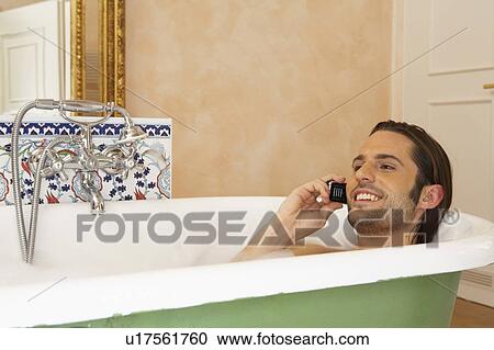 Stock Image - young man smiling in bath holding mobile phone, high section. Fotosearch