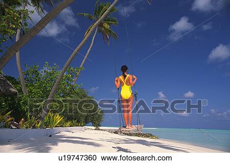 Stock Image -  a Woman in a Bathing Suite, Standing on a Swing Looking at the Ocean, Rear View, Maldives, Micronesia. Fotosearch