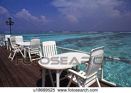 Stock Photography -  Several White Tables and Chairs on a Porch, Right Next to the Clear Water of the Ocean, Side View, Maldives, Micronesia. Fotosearch