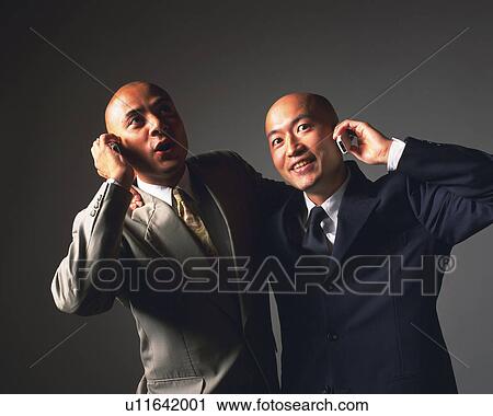 Stock Image - Two bald headed businessmen holding shoulders and talking on mobile phones, Front View. Fotosearch