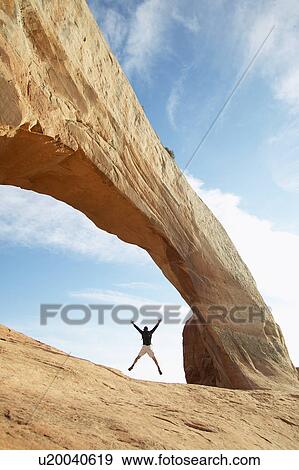 Stock Photo - Man jumping under arched rock. Fotosearch