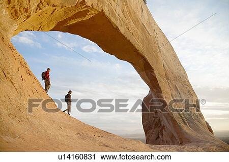 Stock Image - Two hikers walking under arched rock. Fotosearch