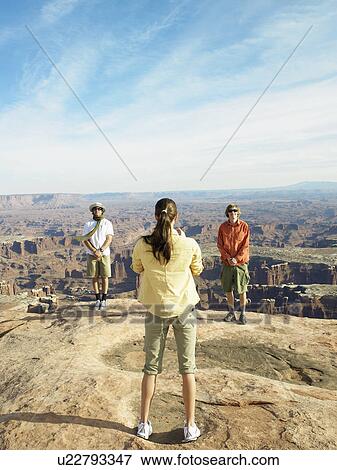 Stock Photo - Woman photographing two men on cliff, rock strata in background. Fotosearch