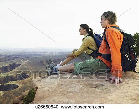 Stock Image - Young couple sitting on cliff and looking at view. Fotosearch