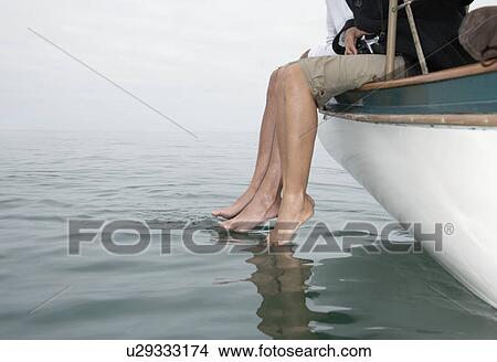 Couple sitting on edge of boat, dipping feet in water (low angle view) View Large Photo Image Picture - Couple sitting on edge of boat, dipping feet in water (low angle view). Fotosearch