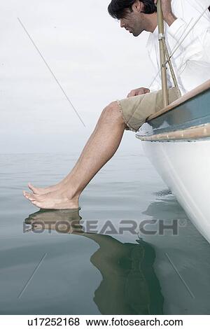 Stock Photo - Man sitting on edge of boat, dipping feet in water (low angle view). Fotosearch