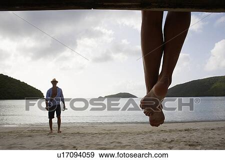 Stock Photo - Mid adult man standing on beach, woman's legs in foreground, St. John, US Virgin Islands, USA. Fotosearch