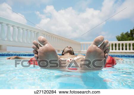 Picture - Young man lying on pool raft in swimming pool (focus on feet, surface level). Fotosearch