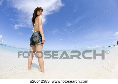 Stock Image - Portrait of a young woman standing on beach, smiling and looking back, fish-eye lens, Saipan, USA . Fotosearch