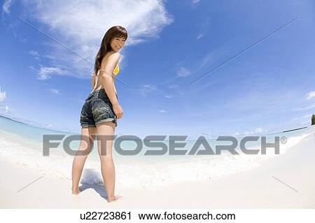 Stock Image - Portrait of a young woman standing on beach, smiling and looking back, fish-eye lens, Saipan, USA . Fotosearch