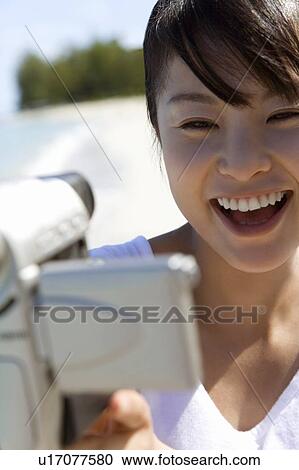 Stock Image - Young woman holding video camera on beach, smiling, differential focus, Saipan, USA . Fotosearch