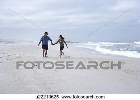 Stock Photography - Mid adult couple running hand in hand on beach, Cape Town, South Africa. Fotosearch