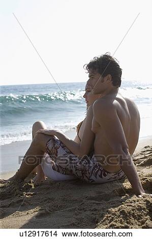 Picture - Young adult couple sitting together on beach, Side View. Fotosearch