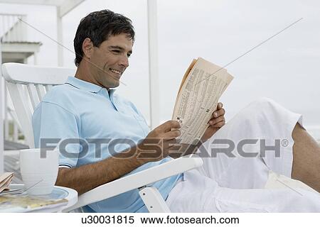 Stock Photography - Mid adult man reading newspaper on lounge chair. Fotosearch