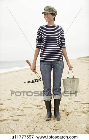 Stock Photo - Woman with pail and pitchfork standing on beach. Fotosearch