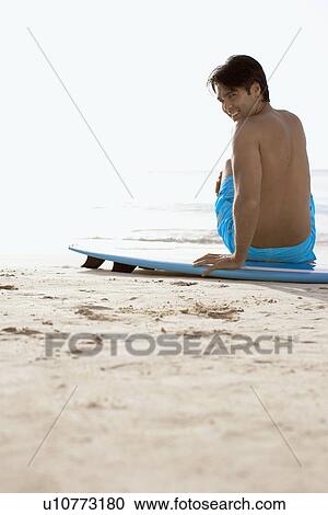 Young man sitting on surfboard on beach View Large Photo Image Stock Image - Young man sitting on surfboard on beach. Fotosearch