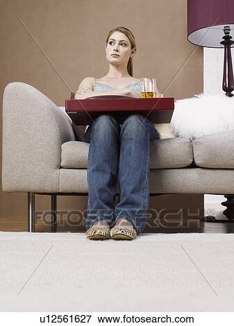 Stock Photo - Young woman with tray sitting on sofa (low angle view). Fotosearch