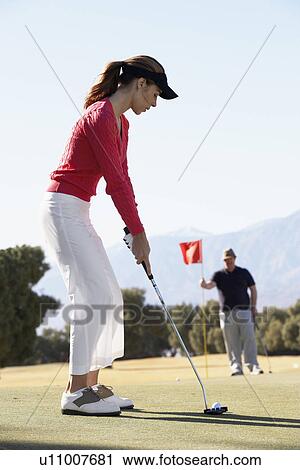 Stock Image - Woman playing golf, man holding golf flag. Fotosearch