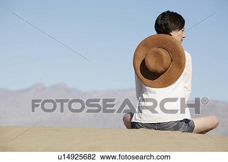 Stock Image - Woman with sun hat sitting on sand dune (rear view). Fotosearch