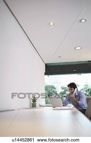 Stock Image - Young businessman using laptop talking on phone (focus on background). Fotosearch