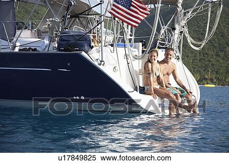 Stock Photography - Mid adult couple sitting at the edge of sailboat. Fotosearch