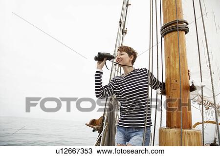 Stock Image - Mid adult woman using binoculars in sailboat. Fotosearch