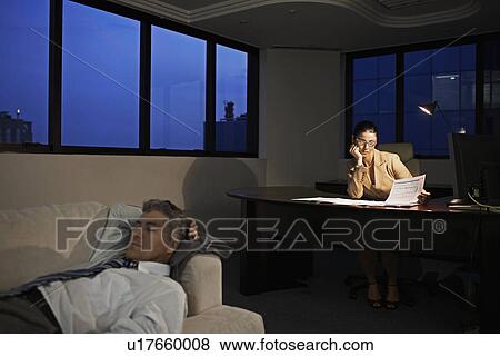 Stock Photo - Businesswoman Working Late, Man Sleeping on Sofa in Office. Fotosearch