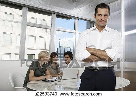 Stock Image - Male manager with colleagues using laptop in background (portrait). Fotosearch