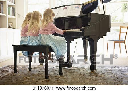 Stock Image - Triplets playing piano in living room (rear view). Fotosearch