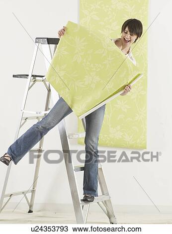 Stock Image - Young Woman Balancing on Stepladder and Holding Wallpaper. Fotosearch