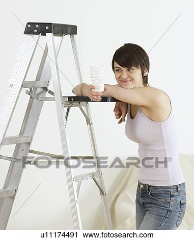 Stock Image - Young Woman Leaning on Stepladder and Holding Light bulb. Fotosearch