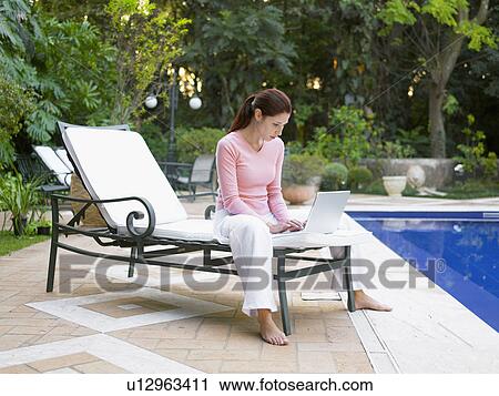 Stock Image - Young woman using laptop by swimming pool. Fotosearch
