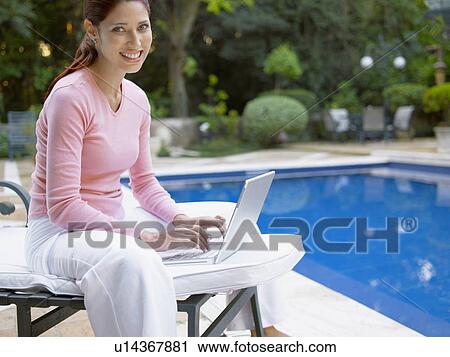 Stock Image - Young woman using laptop by swimming pool (portrait). Fotosearch
