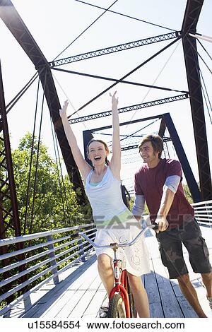 Picture - Woman cycling with raised hands, man holding her bicycle. Fotosearch