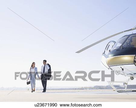 Stock Image - Two businesspeople walking on runway near helicopter. Fotosearch