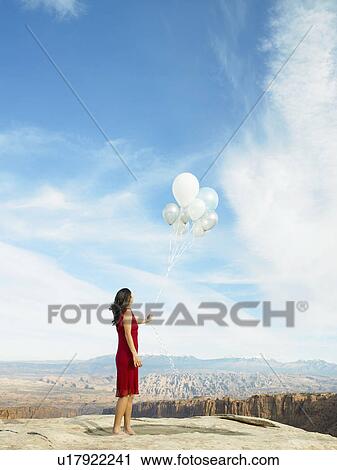 Stock Image - Mid adult woman holding balloons, rock strata in background. Fotosearch