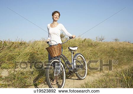 Stock Image - Mid adult woman with bicycle walking on path. Fotosearch