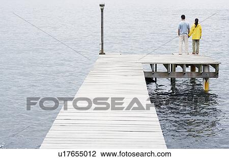 Stock Image - Young couple standing on jetty and looking at view. Fotosearch