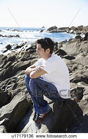 Young man sitting on rock by sea View Large Photo Image Stock Photograph - Young man sitting on rock by sea. Fotosearch