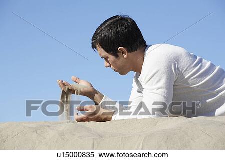 Stock Photography - Man lying on front and sifting sand through hand. Fotosearch