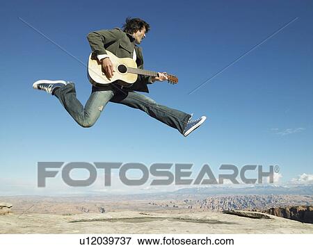 Stock Photo - Mid adult man jumping and playing acoustic guitar, rock strata in background. Fotosearch