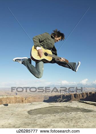 Stock Photo - Mid adult man jumping and playing acoustic guitar, rock strata in background. Fotosearch