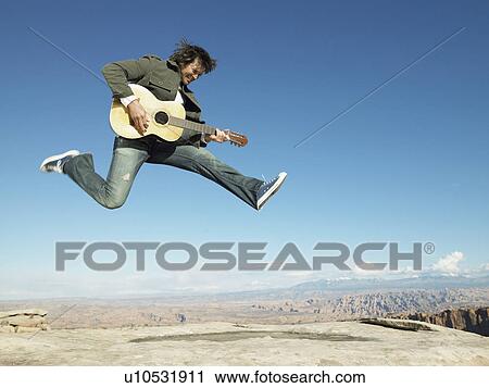Stock Image - Mid adult man jumping and playing acoustic guitar, rock strata in background. Fotosearch