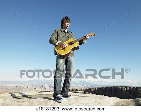 Stock Image - Mid adult man playing acoustic guitar, rock strata in background, low angle view. Fotosearch